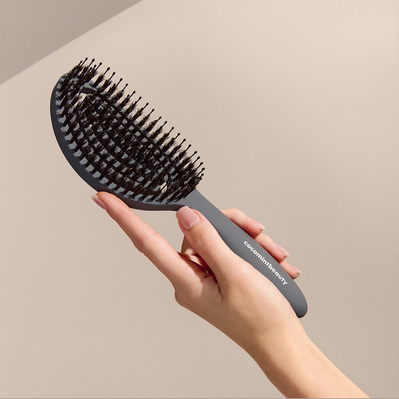 Hand holding a black hairbrush with 'cocomintbeauty' branding against a beige background